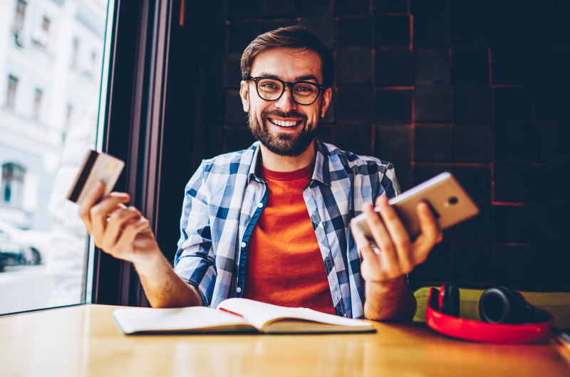 Foto de um homem sorrindo segurando um celular e um cartão de crédito | Pix | Dúvidas sobre dívidas | Eu Dou Conta