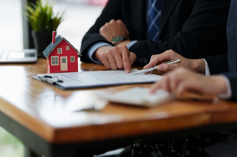 Duas pessoas em uma mesa assinando documentos imobiliários, com uma pequena casa em miniatura ao lado, simbolizando a formalização da compra de um imóvel e a aplicação do ITBI.