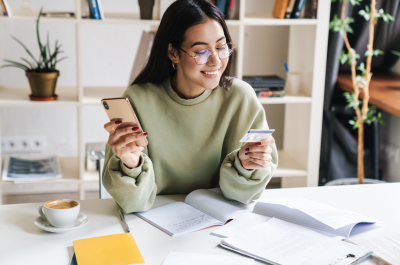 Dicas para evitar dívidas | Jovem mulher sorrindo segurando o celular e um cartão de crédito | Como limpar o seu nome | Eu Dou Conta