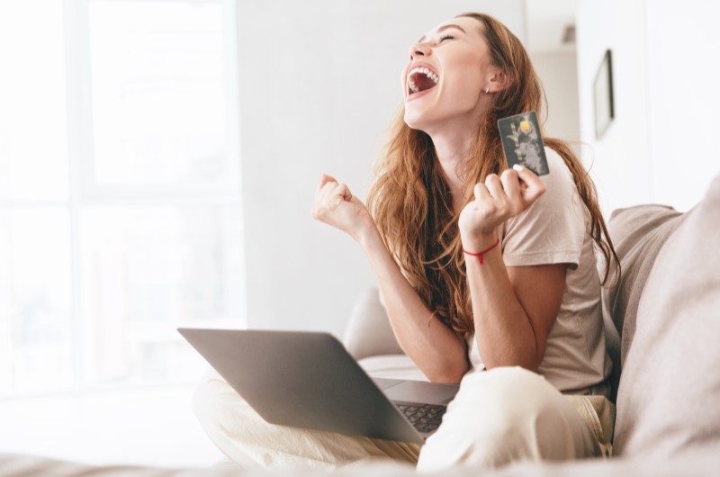 Cartão de crédito | Jovem feliz segurando um cartão de crédito e pesquisando no computador | Como limpar o seu nome | Eu Dou Conta