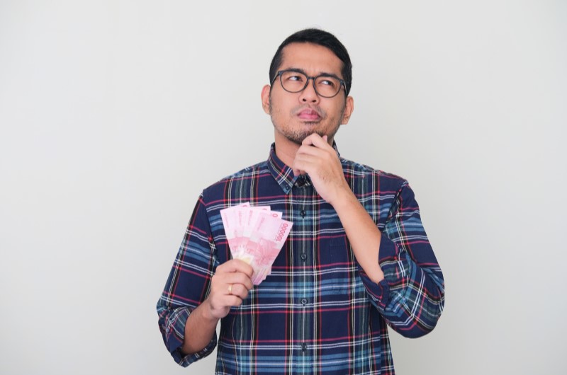 Perguntas frequentes | Foto de um homem segurando notas de dinheiro com cara de dúvida | Economia e renda extra | Eu Dou Conta