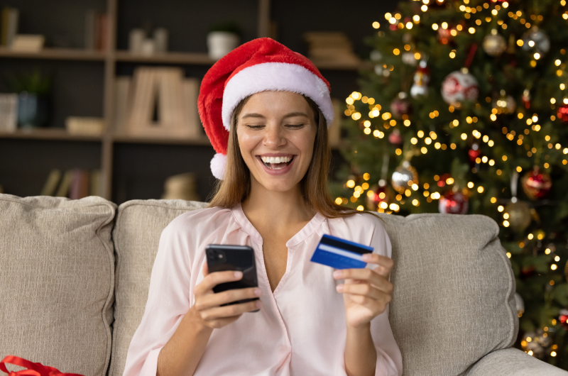 Como evitar o nome negativado nas compras de fim de ano | Foto de uma mulher sorridente segurando o celular e um cartão de crédito fazendo compras para o Natal | Como limpar o seu nome | Eu Dou Conta