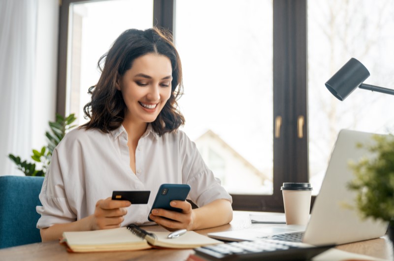 O que é cartão de crédito? | Foto de uma mulher segurando um celular e um cartão de crédito em frente ao notebook | Economia e renda extra | Eu Dou Conta