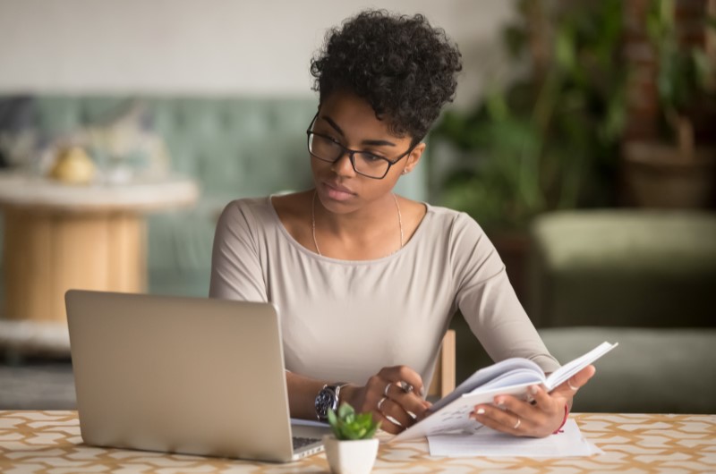 Defina suas metas e objetivos financeiros | Foto de uma moça concentrada enquanto olha para o notebook e segura um caderno | Economia e renda extra | Eu Dou Conta 