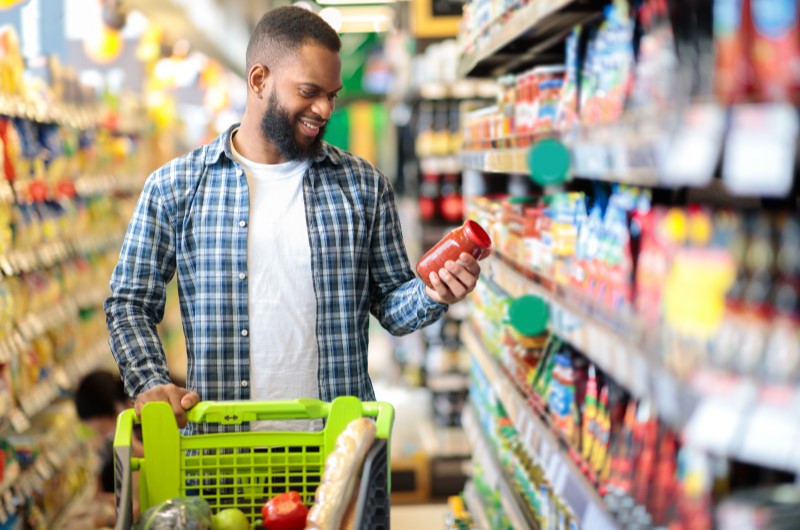 Tenha disciplina na hora de ir às compras | Foto de um homem segurando um alimento enquanto faz compras no supermercado | Eu Dou Conta | Blog da Tenda