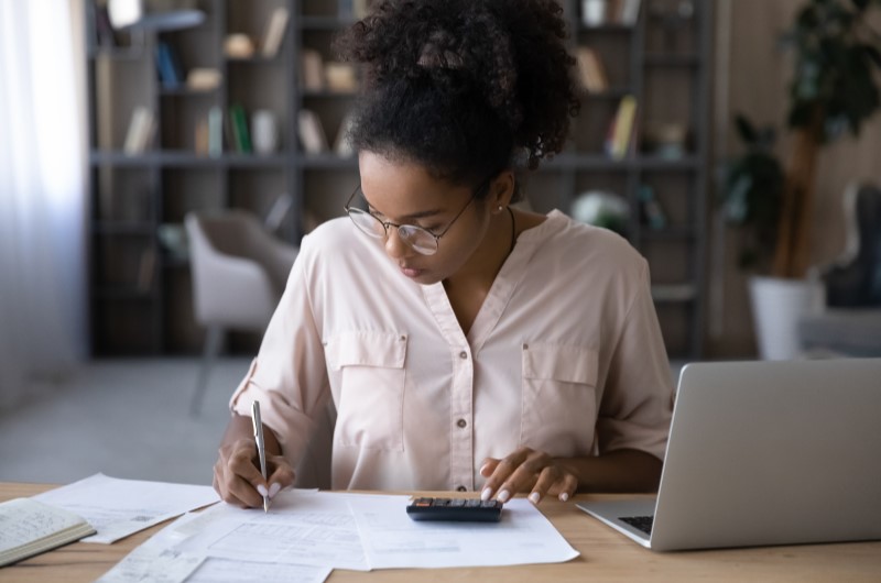 Dicas para reduzir as despesas variáveis | Foto de um cofrinho, moedas, papéis e calculadora sobre uma mesa | Economia e renda extra | Eu Dou Conta 