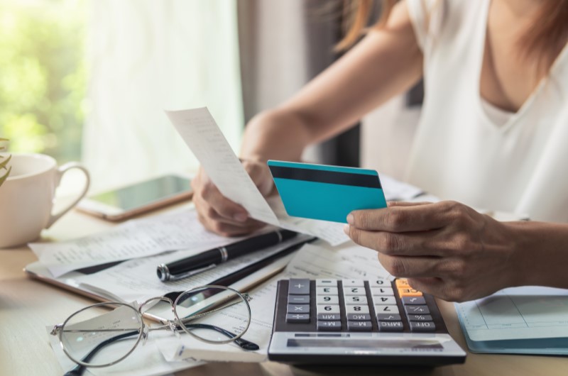 Como saber se estou com cartão inadimplente? | Foto de uma mulher segurando um cartão e um papel, com um óculos e calculadora sobre a mesa | Dúvidas sobre dívidas | Eu Dou Conta 