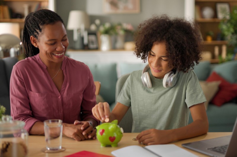 Maior economia das finanças | Foto de uma mãe sorridente, observando o filho colocar moedas em um cofrinho | Economia e Renda Extra | Eu Dou Conta 