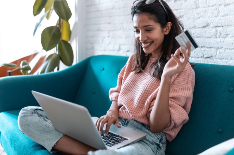 Quitou as dívidas? O que fazer para não voltar a ter um cartão inadimplente? | Foto de uma moça sorridente, segurando um cartão e usando o notebook | Dúvidas sobre dívidas | Eu Dou Conta 