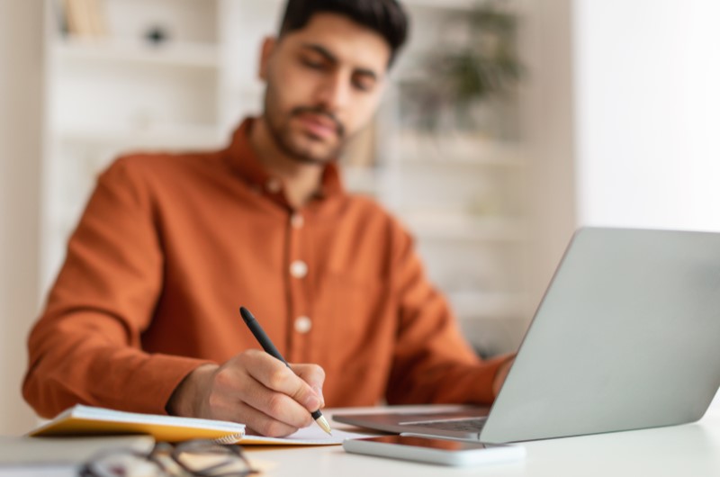 Defina objetivos | Foto de um homem usando o notebook e escrevendo em um caderno | Economia e renda extra | Eu Dou Conta 