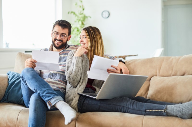 Vantagens de antecipar o pagamento do financiamento | Foto de um casal no sofá, usando o notebook e segurando alguns papeis | Dúvidas sobre dívidas | Eu Dou Conta 