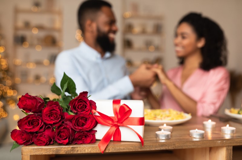 8 ideias para aproveitar o Dia dos Namorados em casa | Foto de um buque de rosas e um presente ao lado, com um casal de namorados ao fundo | Casa e decoração | Blog da Tenda