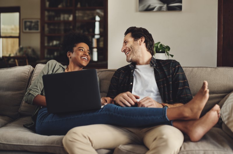 Lazer | Foto de um casal sentado no sofa enquanto usam o notebook e sorriem | Trilha da Conquista | Blog da Tenda