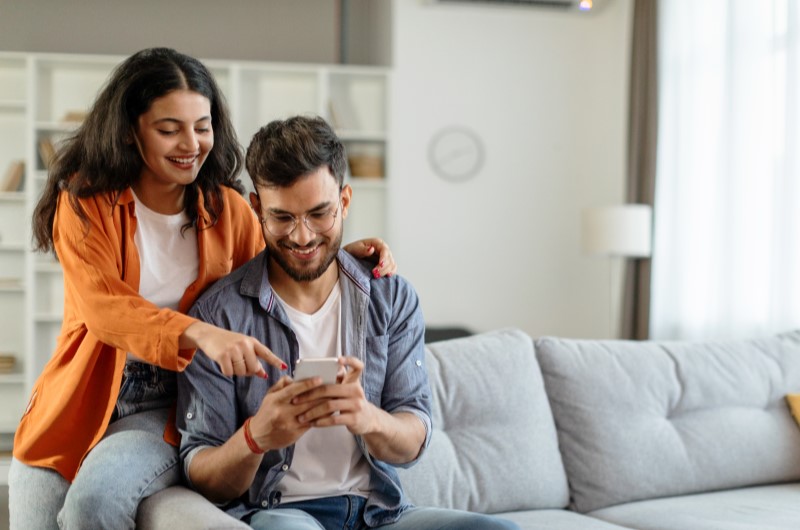 O que fazer no bairro Taquara | Foto de um casal sorridente olhando o celular na sala de estar | Trilha da Conquista | Blog da Tenda