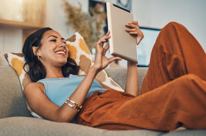 Onde fica o bairro Taquara | Foto de uma moça deitado no sofá, sorrindo enquanto usa o tablet | Trilha da Conquista | Blog da Tenda