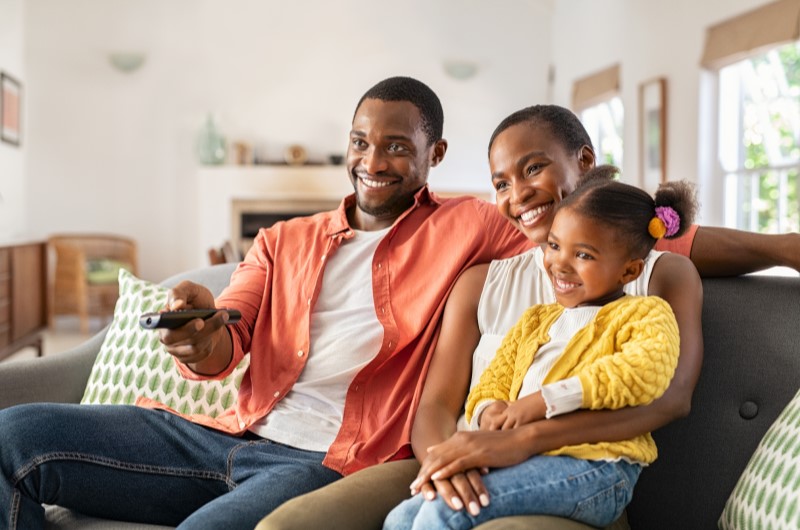 5 vantagens de morar em condomínio fechado | Foto de um casal casal sorridente com uma filha no sofá da sala de estar | Viver em condomínio | Blog da Tenda