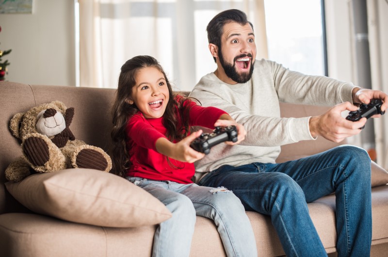 8 ideias para aproveitar as férias escolares em casa | Foto de um pai e uma filha sorridentes, jogando video game na sala de estar | Viver em condomínio | Blog da Tenda