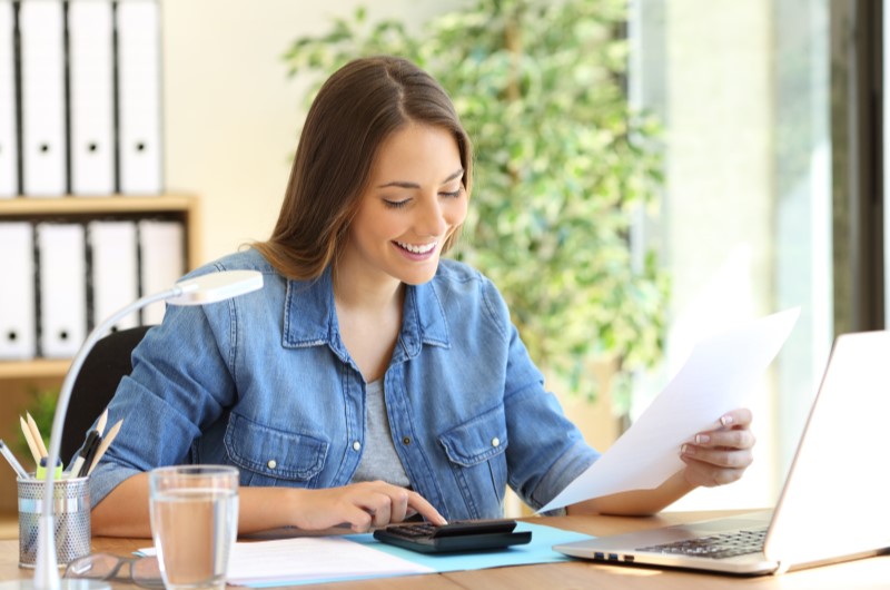 Alcance de metas | Foto de uma moça sorrindo enquanto olha para um papel e usa a calculadora | Economia e renda extra | Eu Dou Conta