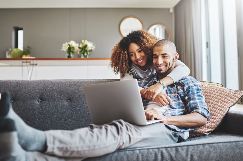 Categoria celebrar | Foto de um casal sorridente no sofá, enquanto olha para o notebook | Trilha da Conquista | Blog da Tenda