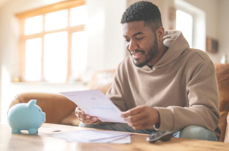 Como organizar as finanças e sair do vermelho? | Foto de um homem segurando um papel com um cofrinho ao lado | Economia e renda extra | Eu Dou Conta 