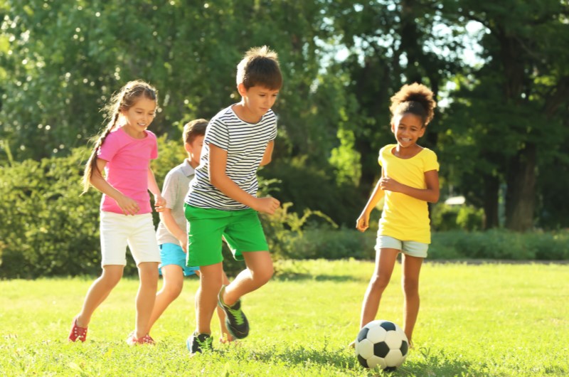 Dicas de passeios para fazer nas férias escolares | Foto de um grupo de crianças jogando bola em um dia ensolarado no parque | Viver em condomínio | Blog da Tenda