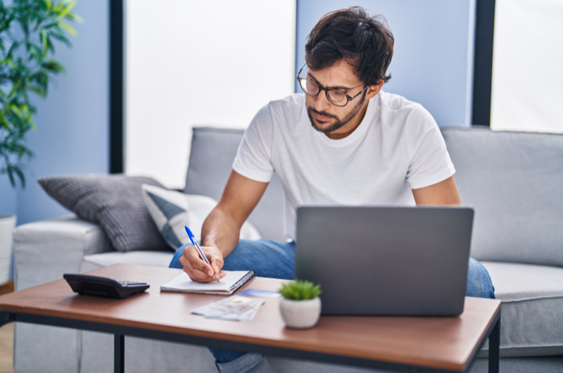 Não assuma novas dívidas | Foto de um homem escrevendo em um caderno e usando um notebook | Como limpar o seu nome | Eu Dou Conta 