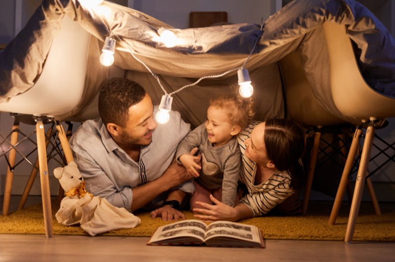 Planeje atividades nas férias escolares | Foto de um pai, mãe e uma criança lendo um livro dentro de uma cabana improvisada | Viver em condomínio | Blog da Tenda