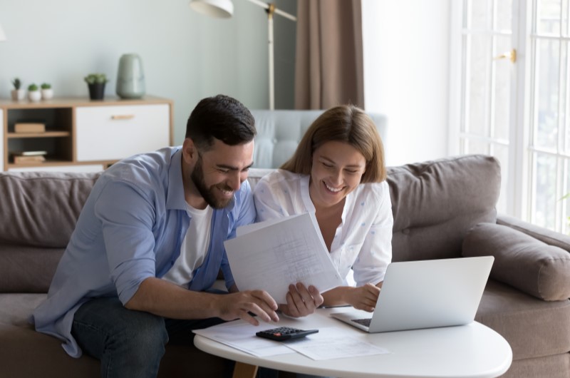 Vantagens de ter independência financeira | Foto de um casal sorridente fazendo contas com uma calculadora e notebook | Economia e renda extra | Eu Dou Conta 