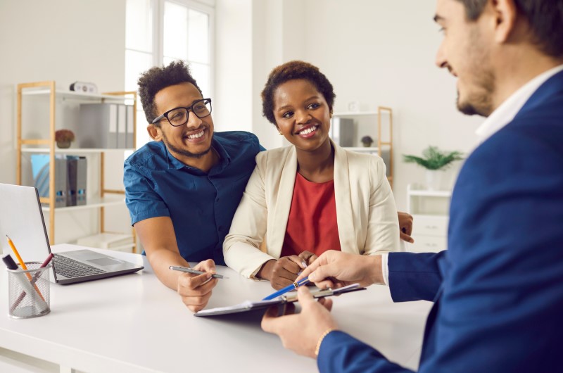 Vantagens e desvantagens do crédito imobiliário | Foto de um casal sorridente conversando com um corretor imobiliário | Trilha da Conquista | Blog da Tenda