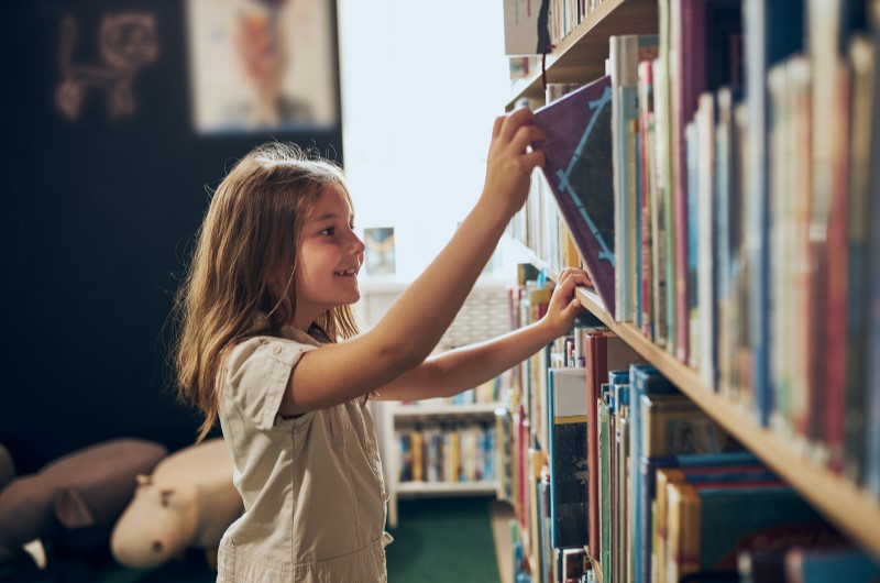 Visite bibliotecas da região | Foto de uma menina pegando um livro em uma biblioteca | Viver em condomínio | Blog da Tenda