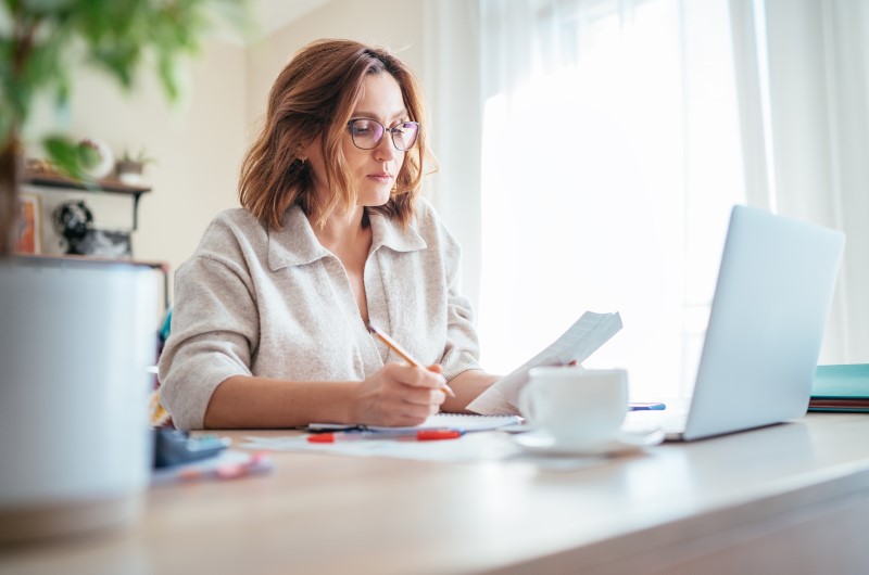 Como evitar contas atrasadas | Foto de uma mulher segurando um lápis e um papel enquanto olha para o notebook | Como limpar o seu nome | Eu Dou Conta 