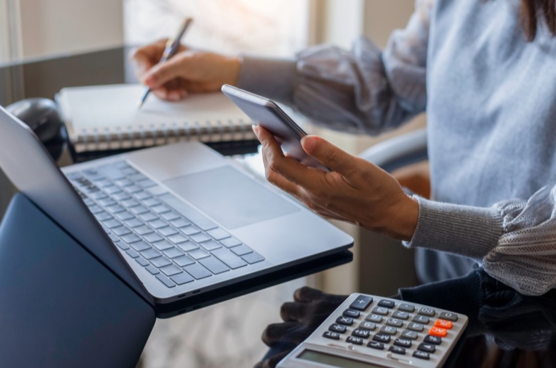 Não ter outros financiamentos ativos | Foto de uma mulher escrevendo em um caderno enquanto usa o celular e notebook | Trilha da Conquista | Blog da Tenda 