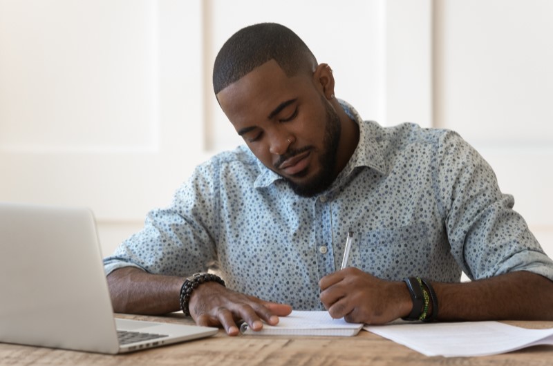 Requisitos para usar o FGTS para comprar imóvel | Foto de um homem escrevendo em um caderno, com um notebook a sua frente | Trilha da Conquista | Blog da Tenda