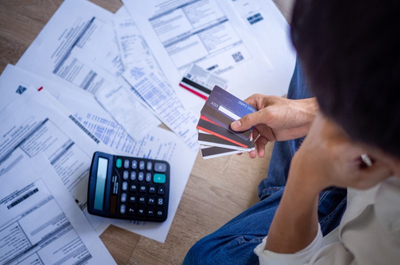 Tenha cuidado com o cartão de crédito | Foto de uma pessoa segurando doversos cartões e olhando para uma calculadora e contas atrasadas | Como limpar o seu nome | Eu Dou Conta 