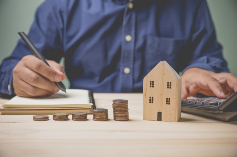 Tipos de imóveis que podem ser comprados com o uso do FGTS | Foto de um homem escrevendo no caderno, com algumas moedas e uma casinha sobre a mesa | Trilha da Conquista | Blog da Tenda