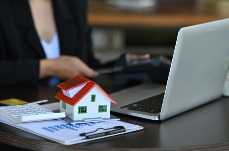 Crédito para reforma e construção | Notebook, casinha em miniatura e calculadora sobre a mesa com uma mulher ao fundo | Dúvidas sobre dívidas | Eu Dou Conta 