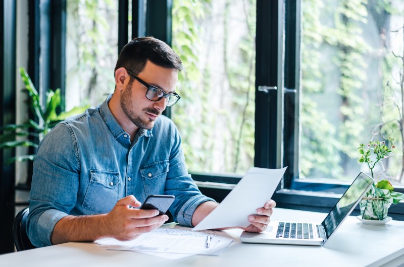 Se planeje com antecedência | Foto de um homem usando o celular e segurando um papel | Economia e renda extra | Eu Dou Conta 