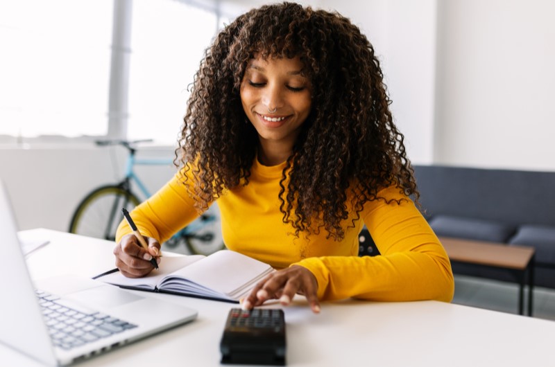 Mantenha um registro e controle das despesas | Foto de uma mulher escrevendo em um caderno enquanto usa a calculadora | Economia e renda extra | Eu Dou Conta