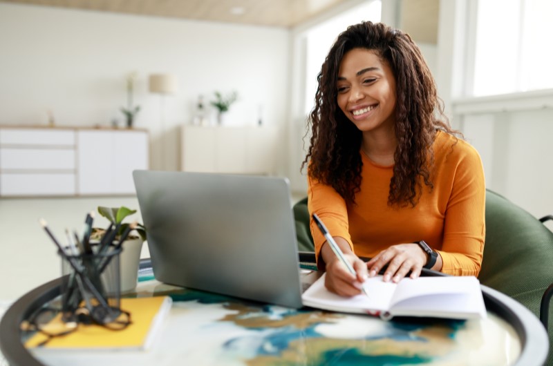 Onde fica o bairro Bonsucesso? | Mulher sorridente escrevendo em um caderno enquanto usa um notebook | Trilha da Conquista | Blog da Tenda