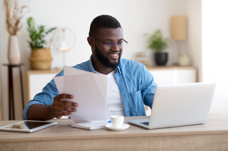 Principais custos ao morar sozinho? | Foto de um homem sorrindo enquanto usa o notebook e segura alguns papeis | Economia e renda extra | Eu Dou Conta