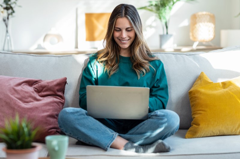 Quais são as vantagens de morar sozinho? | Foto de uma mulher sorrindo no sofá da sala, enquanto usa o notebook | Economia e renda extra | Eu Dou Conta 