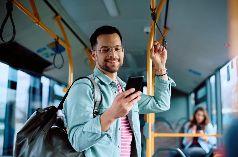 Transporte | Homem sorridente de óculos usando celular em um ônibus | Trilha da Conquista | Blog da Tenda