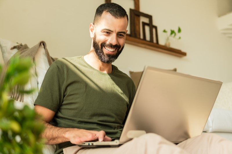 Apartamento em Guaratiba | Homem sorridente usando o notebook na sala de casa | Trilha da Conquista | Blog da Tenda