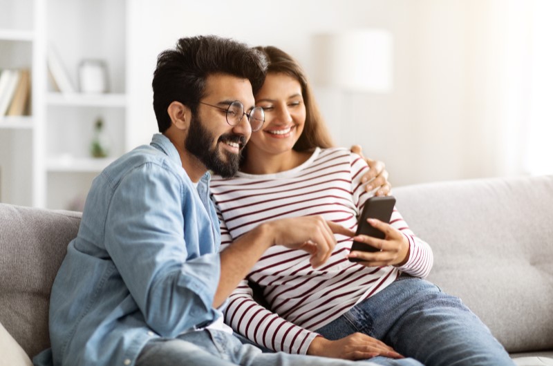 Caxangá | Casal sorridente, sentado no sofá enquanto usando o celular | Trilha da Conquista | Blog da Tenda