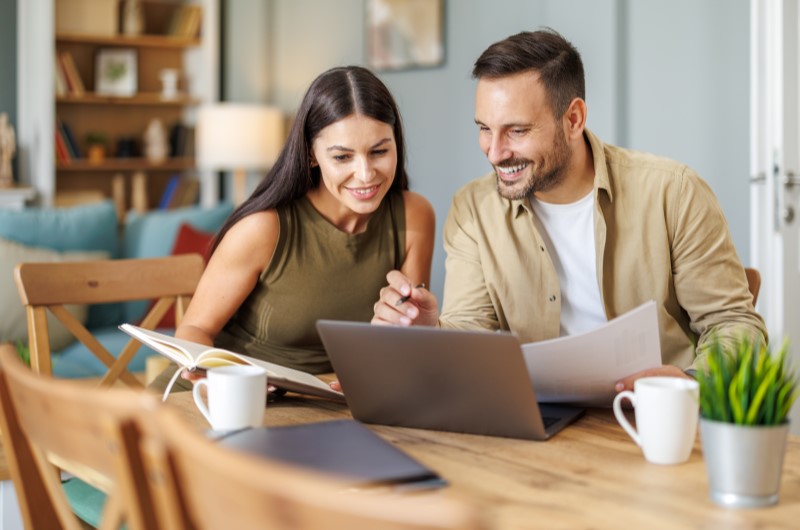 Importância do planejamento financeiro para a vida do casal | Casal sorridente usando o notebook para organizar as finanças | Economia e renda | Eu Dou Conta