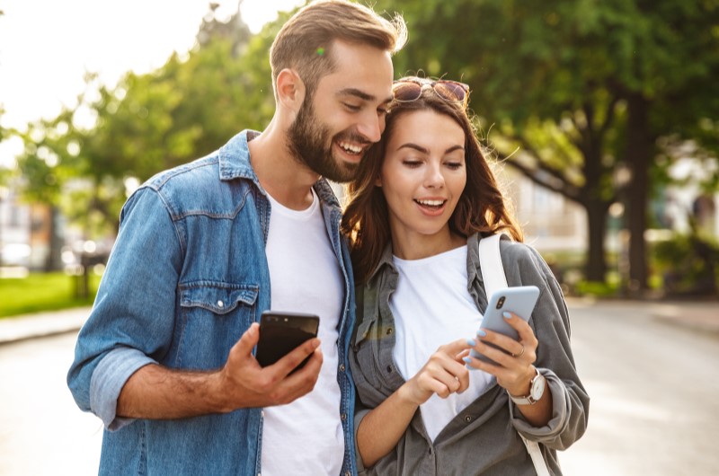 Jacarepaguá | Casal feliz andando em um parque enquanto olham para um celular | Trilha da Conquista | Blog da Tenda 