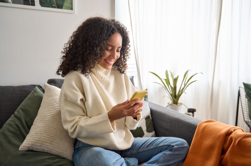 Serviços e comércios | Mulher sorridente usando o celular na sala | Trilha da Conquista | Blog da Tenda