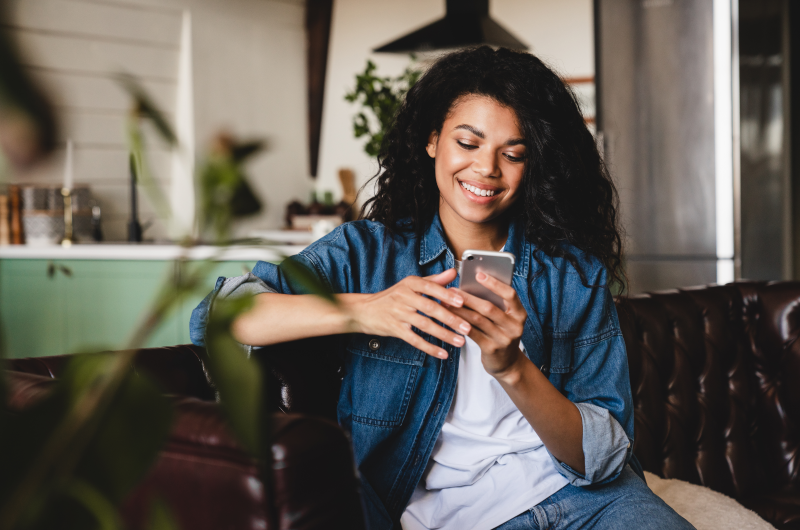 Comércios e serviços | Mulher sorridente usando o celular no sofá da sala | Trilha da Conquista | Blog da Tenda 