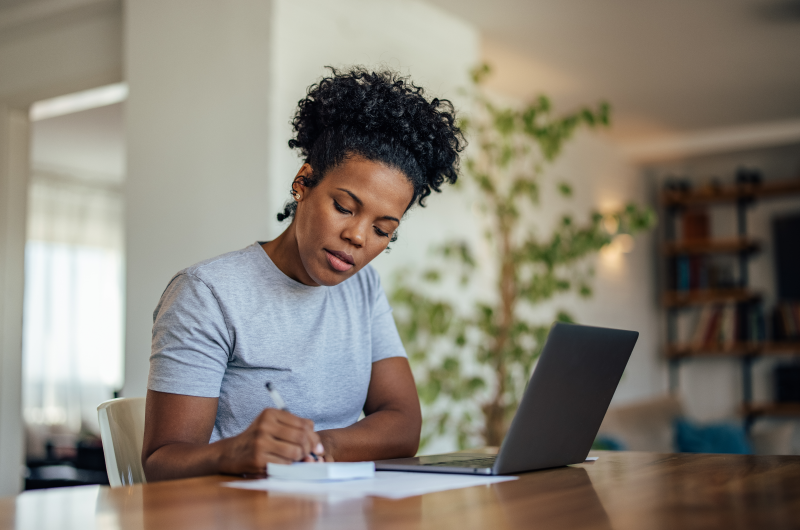 Financiar imóvel apenas para residência | Mulher concentrada em escrever em um bloco de notas e usar o notebook | Blog da Tenda