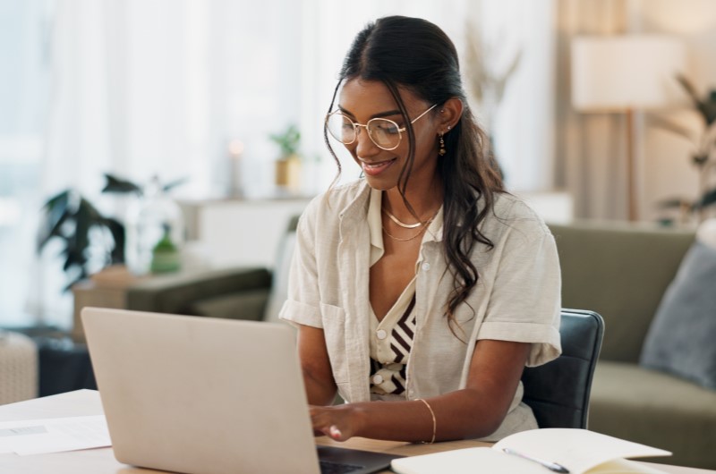 Verifique se você atende aos requisitos | Mulher sorrindo enquanto trabalha em laptop, em ambiente doméstico moderno | Minha Casa Minha Vida | Blog da Tenda 